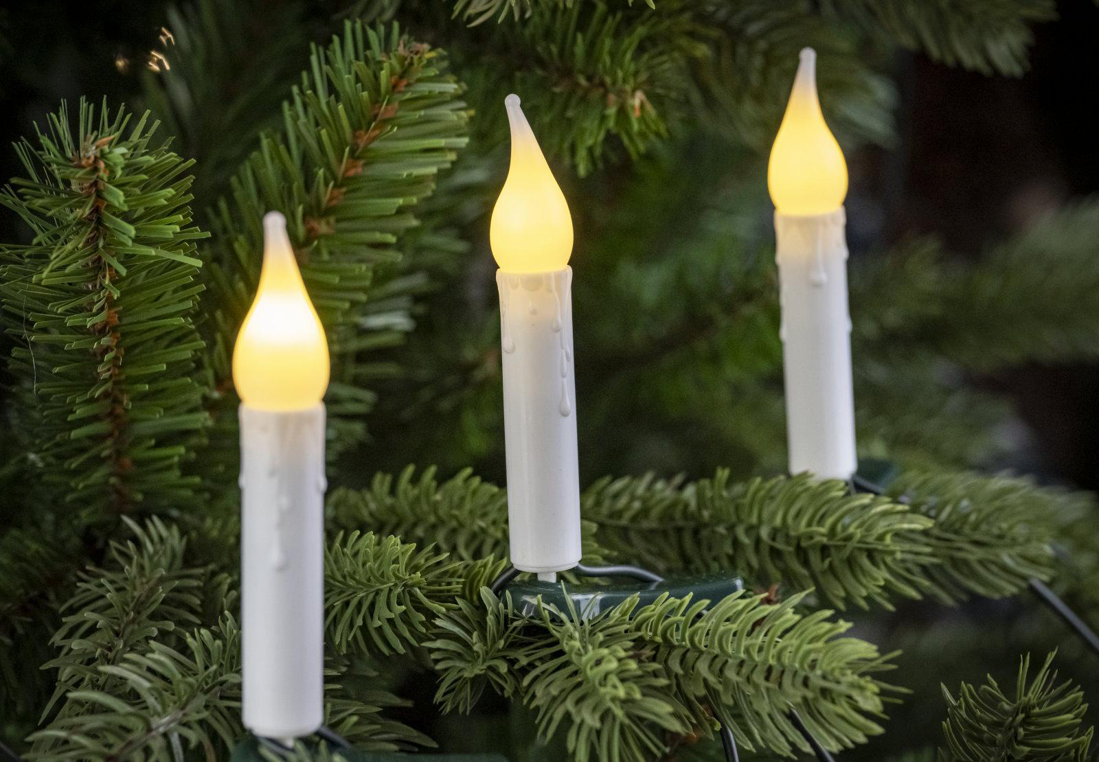 Drei leuchtende, weiße Kerzen auf einem Weihnachtsbaum. Die Kerzenflammen sind gelb. Grüne Nadeln umgeben die Kerzen.