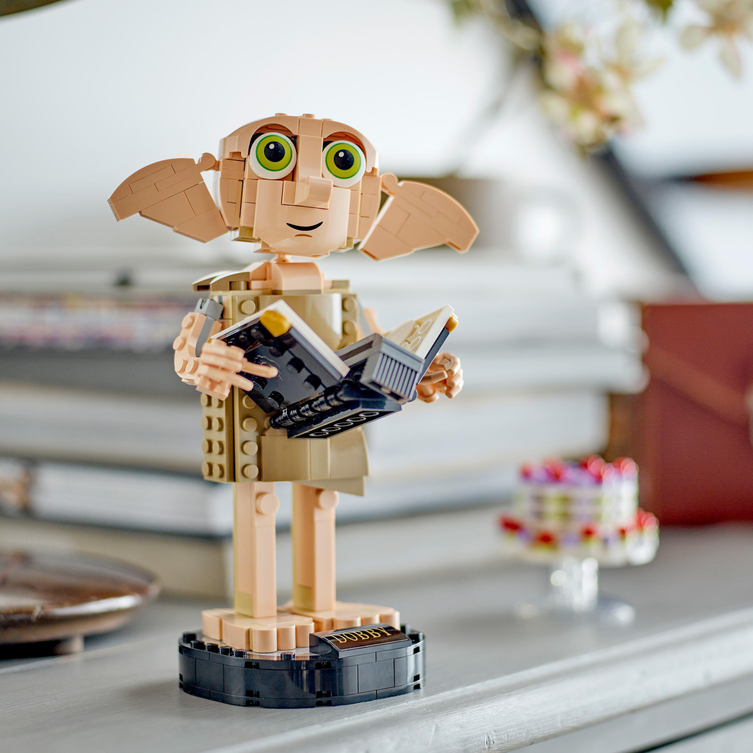 Lego Dobby Figur, hält ein Buch. Im Hintergrund Bücher und ein kleiner Kuchen. Auf hellgrauer Oberfläche.