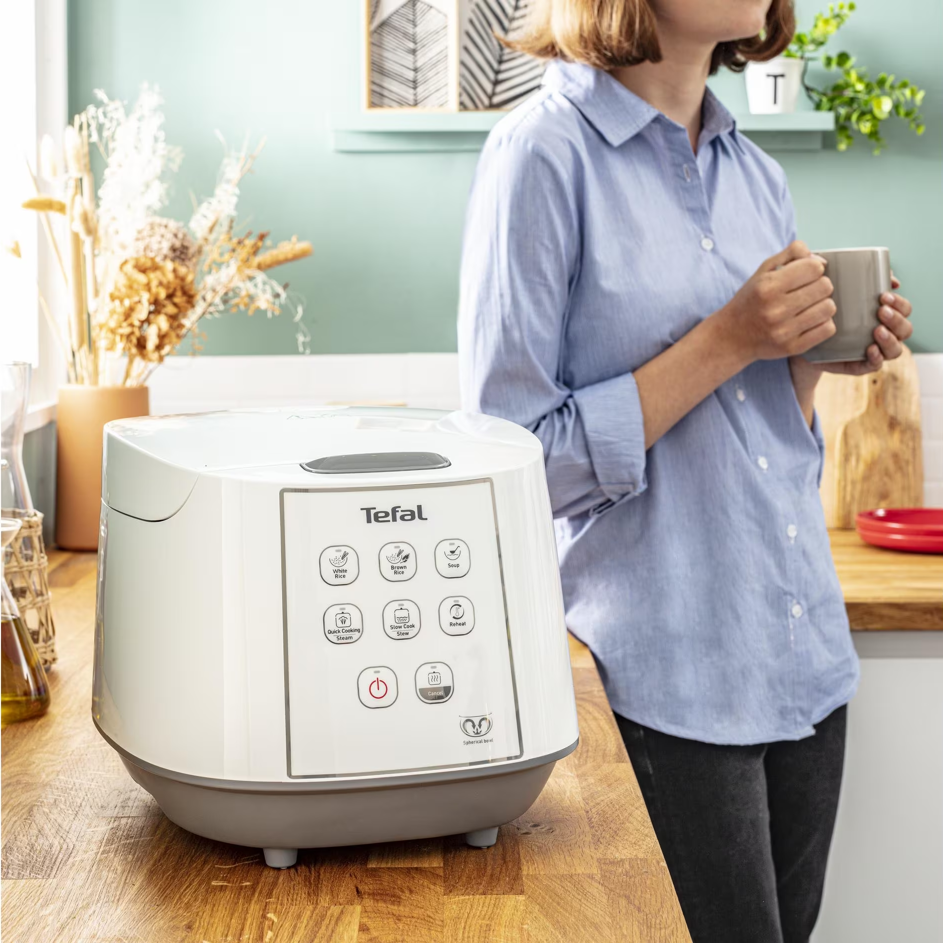 Ein weißer Tefal Reiskocher auf einer Holztheke, mit einer Frau daneben, die einen Becher hält.