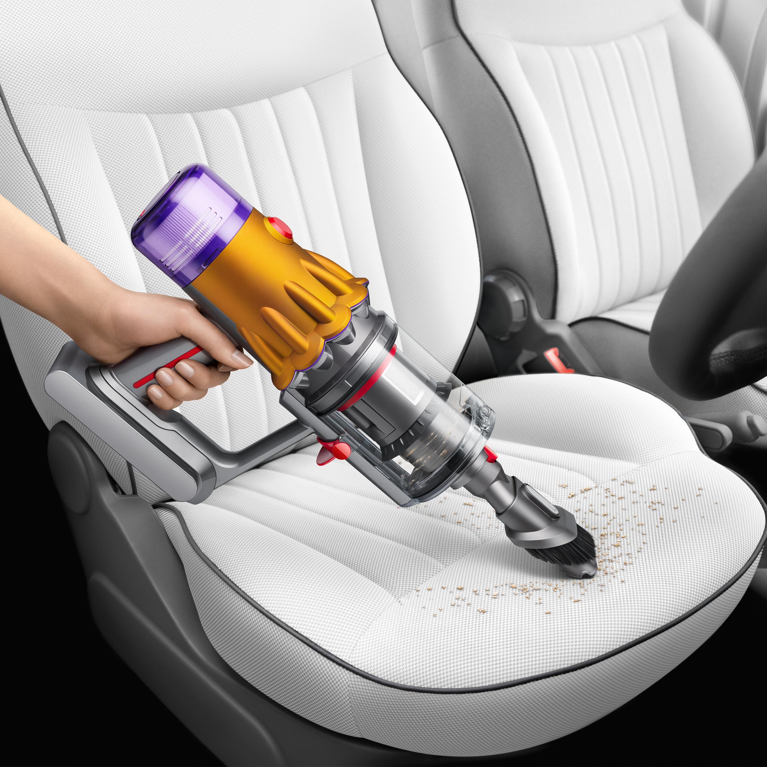 Eine Hand benutzt einen gelb-silbernen Staubsauger auf einem weißen Autositz mit Krümeln.