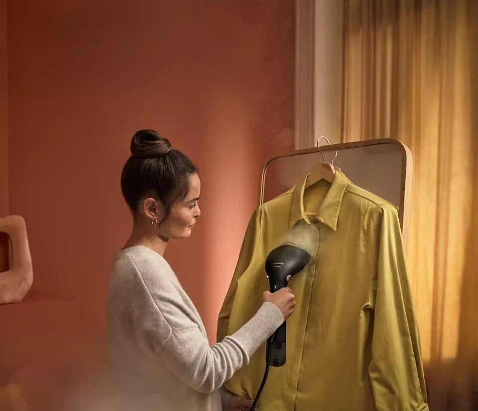 Kobieta prasuje żółtą koszulę wiszącą na lustrze. Różowa ściana w tle. Parownica jest czarna.