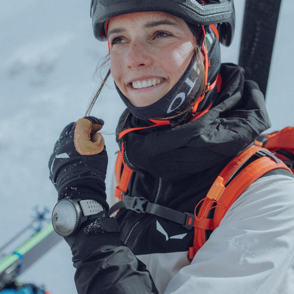 Eine Person in Winterkleidung lächelt, mit Gesichtsbedeckung und Brille, in verschneiter Umgebung.