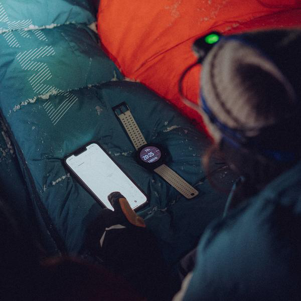 Eine Smartwatch und ein Telefon auf einem Schlafsack. Die Uhr zeigt die Zeit und das Telefon eine Karte.