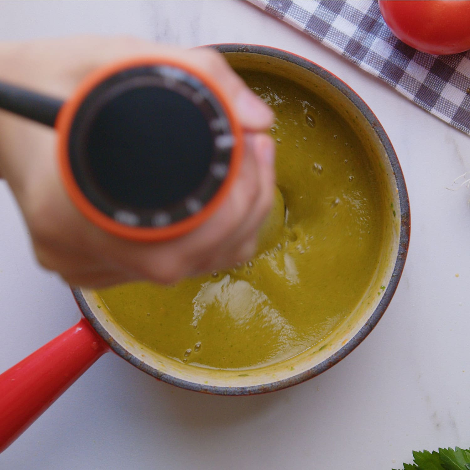 Mano mezclando sopa verde en una olla roja con tomate y perejil, sobre superficie blanca.
