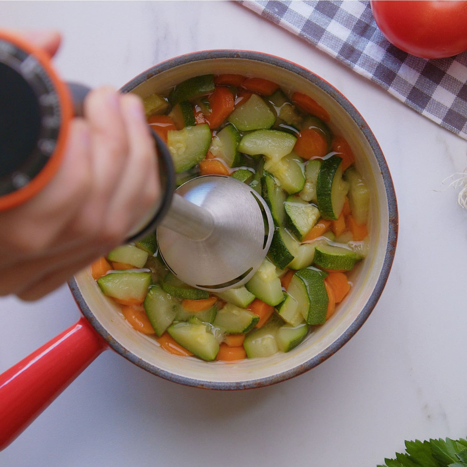Mano usando una licuadora en una olla roja con verduras picadas, preparando sopa. Encimera blanca.