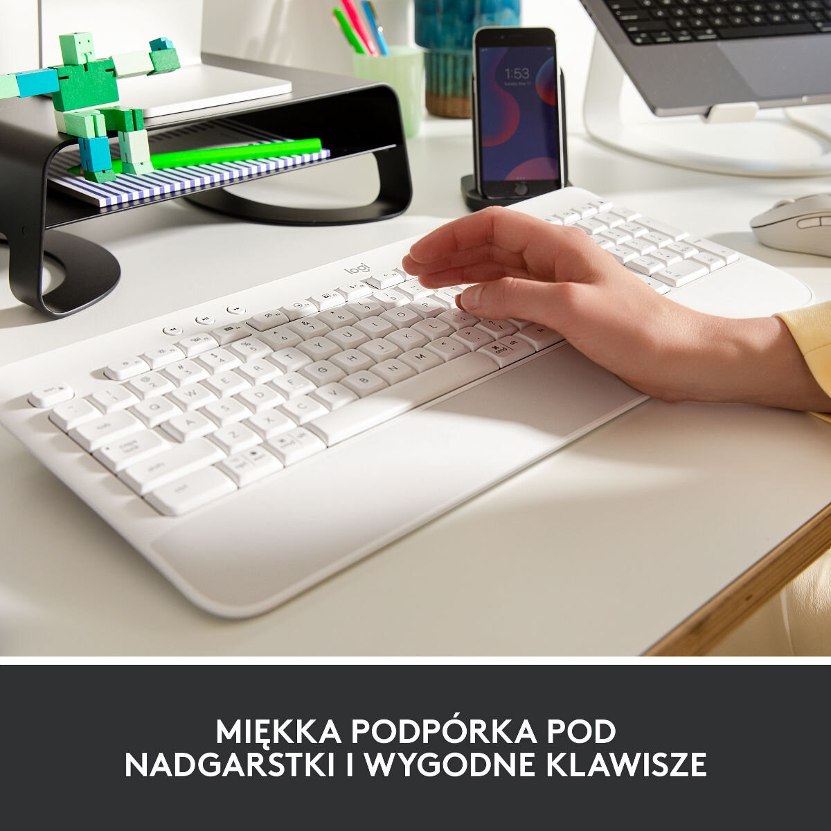 Osoba pisząca na białej klawiaturze z podpórką pod nadgarstek. Telefon i monitor w tle.