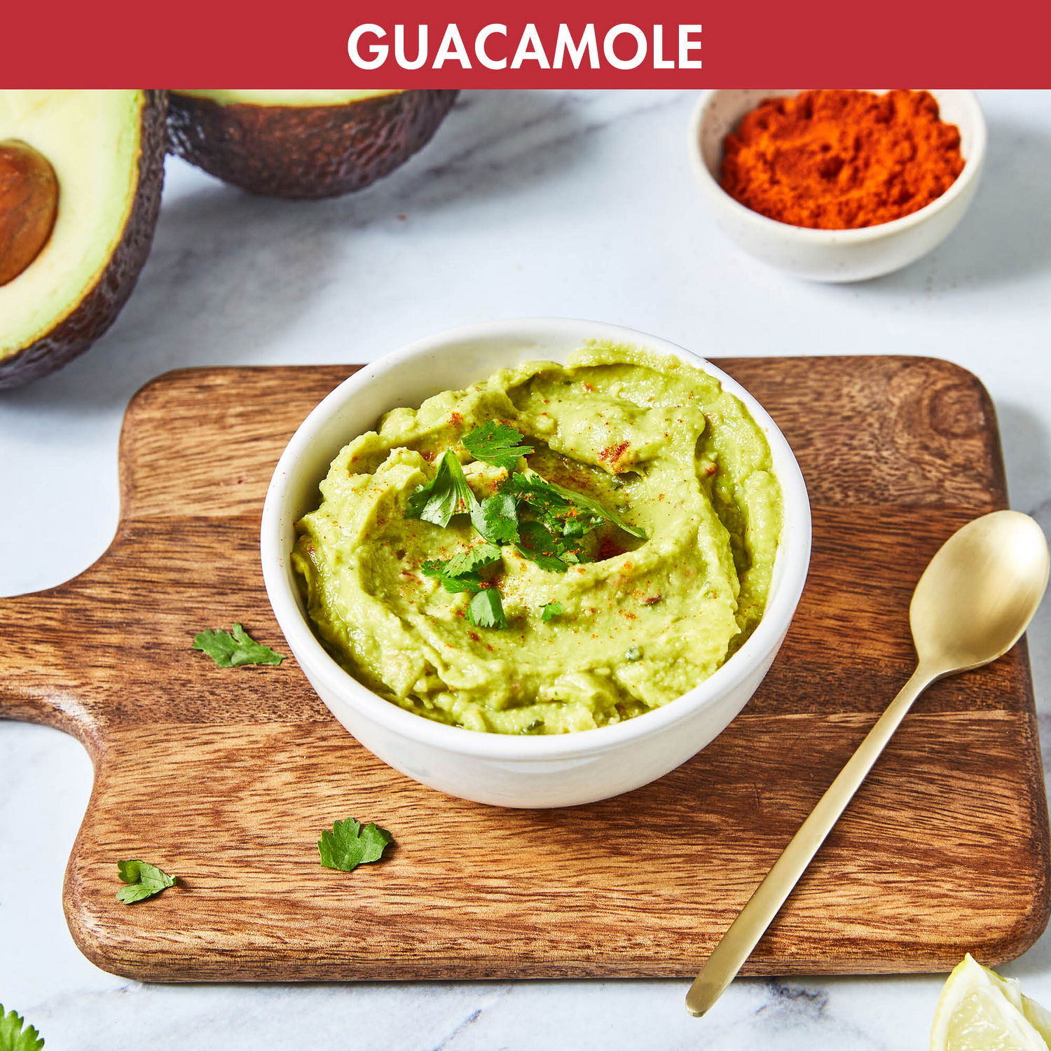 Guacamole in a white bowl on a wooden board. Ingredients and a spoon are on the side.