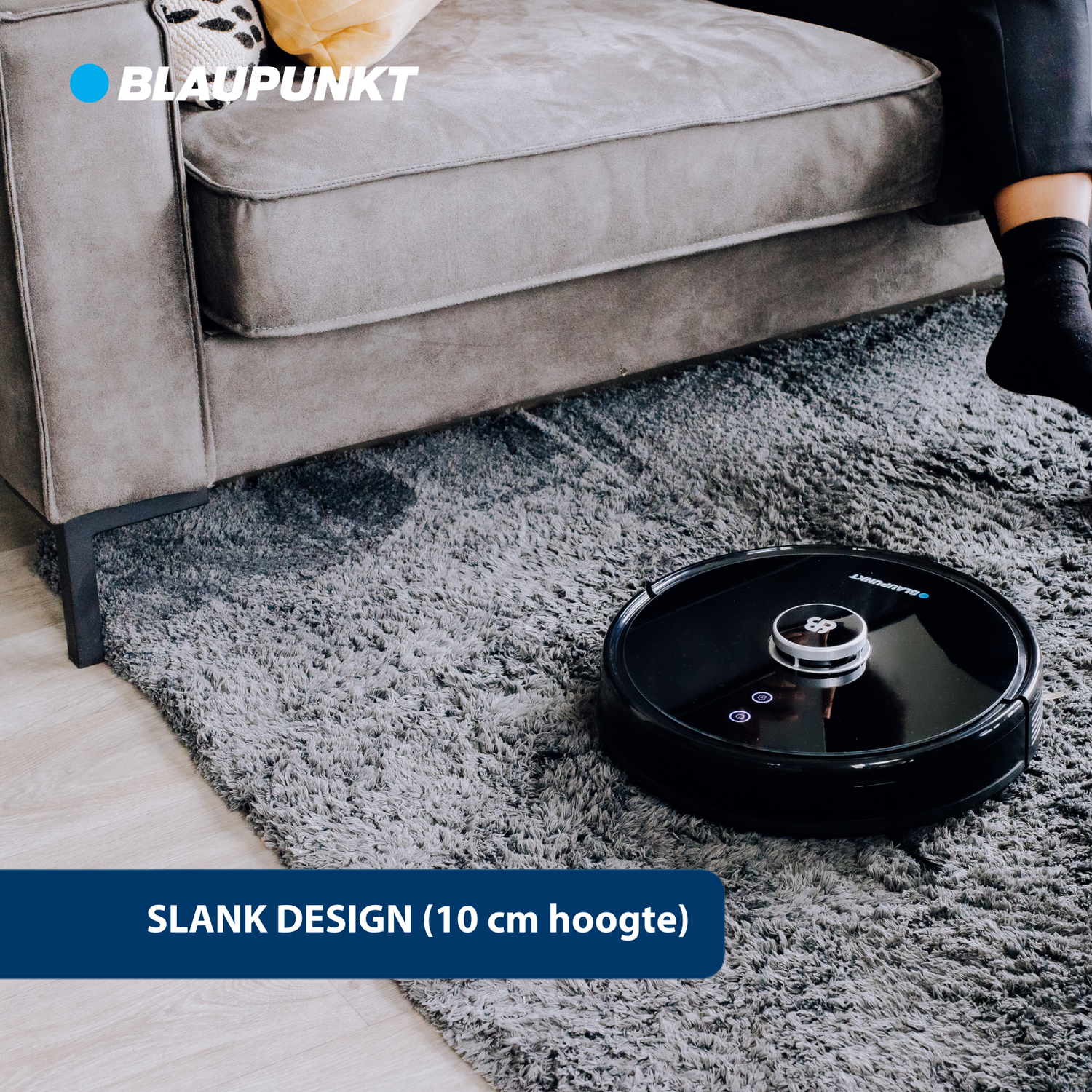 A black robot vacuum cleaner on a gray carpet. A person's legs are in the background.