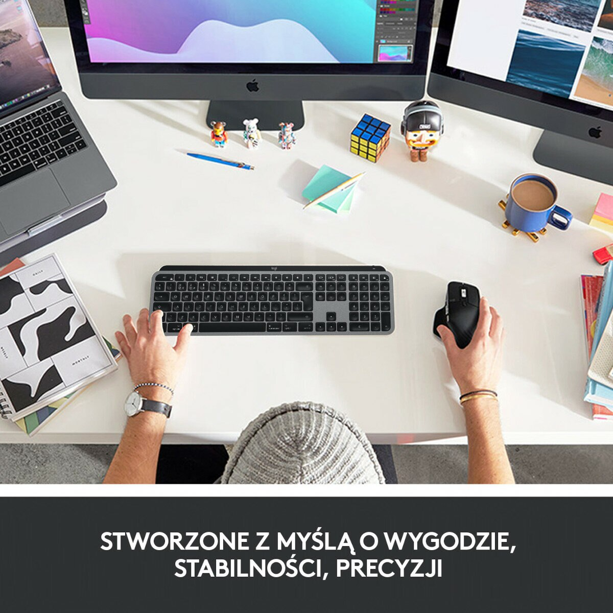Osoba używa klawiatury i myszy na białym biurku. Widoczny monitor, laptop i inne przedmioty.