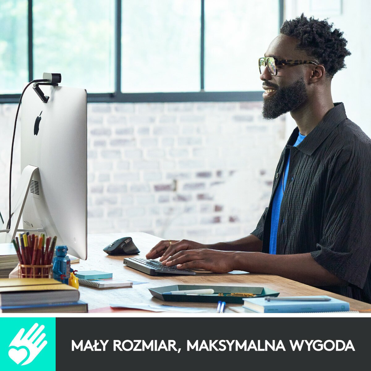 Mężczyzna w okularach używa klawiatury i komputera. Siedzi przy biurku. Kamera internetowa jest na monitorze.