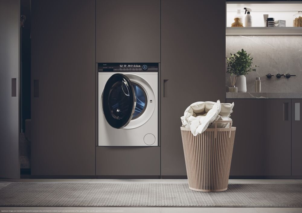 Pralka w pokoju. Kosz z praniem leży na podłodze. Ciemne szafki w tle.
