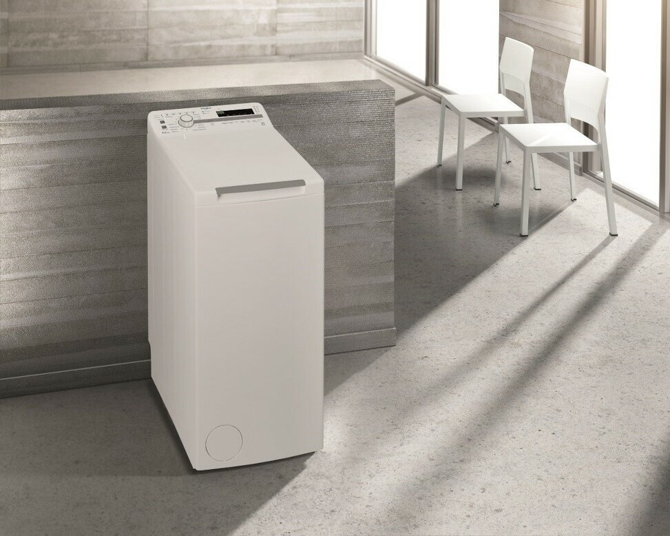 Biała pralka ładowana od góry obok dwóch białych krzeseł, w nowoczesnym pomieszczeniu.
