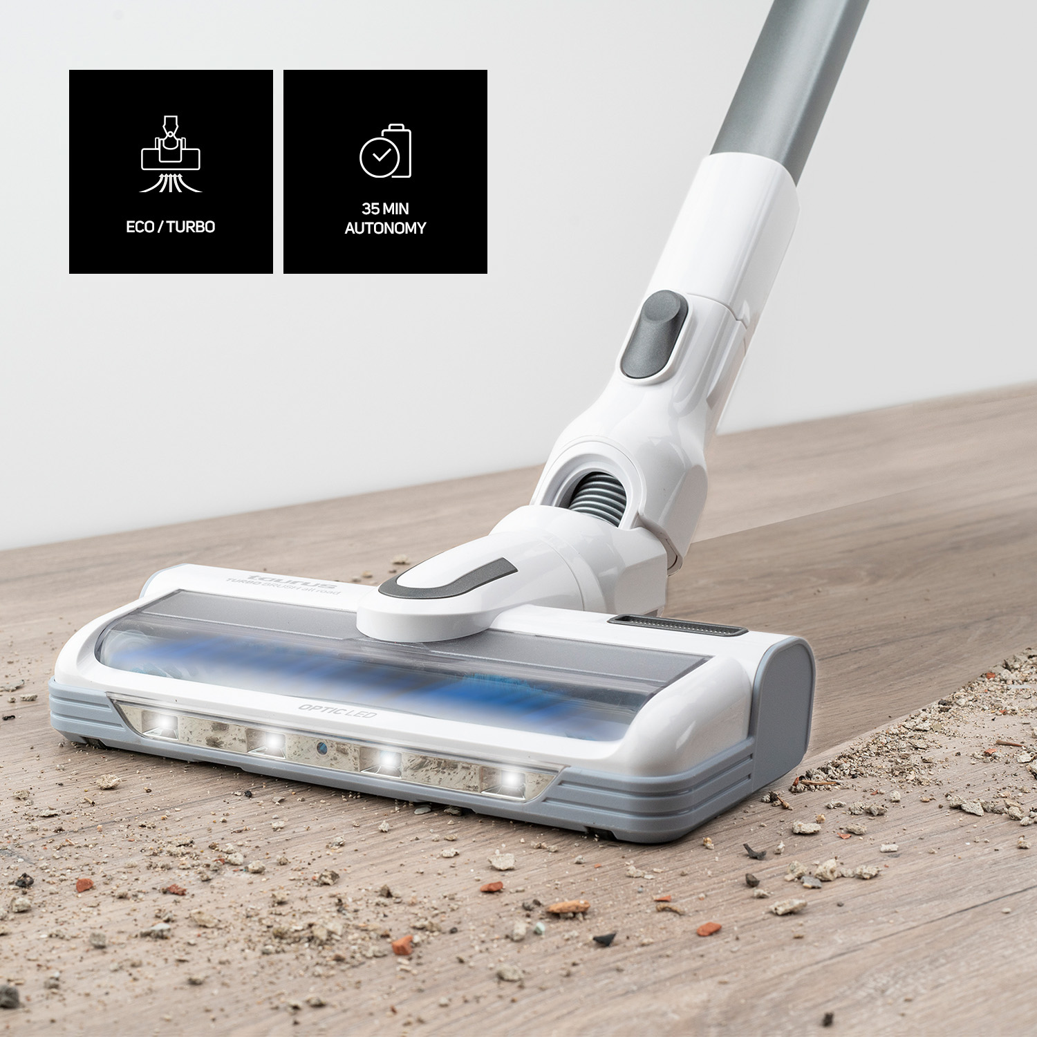 Una aspiradora inalámbrica blanca y gris en un suelo de madera con suciedad, iconos eco/turbo visibles.
