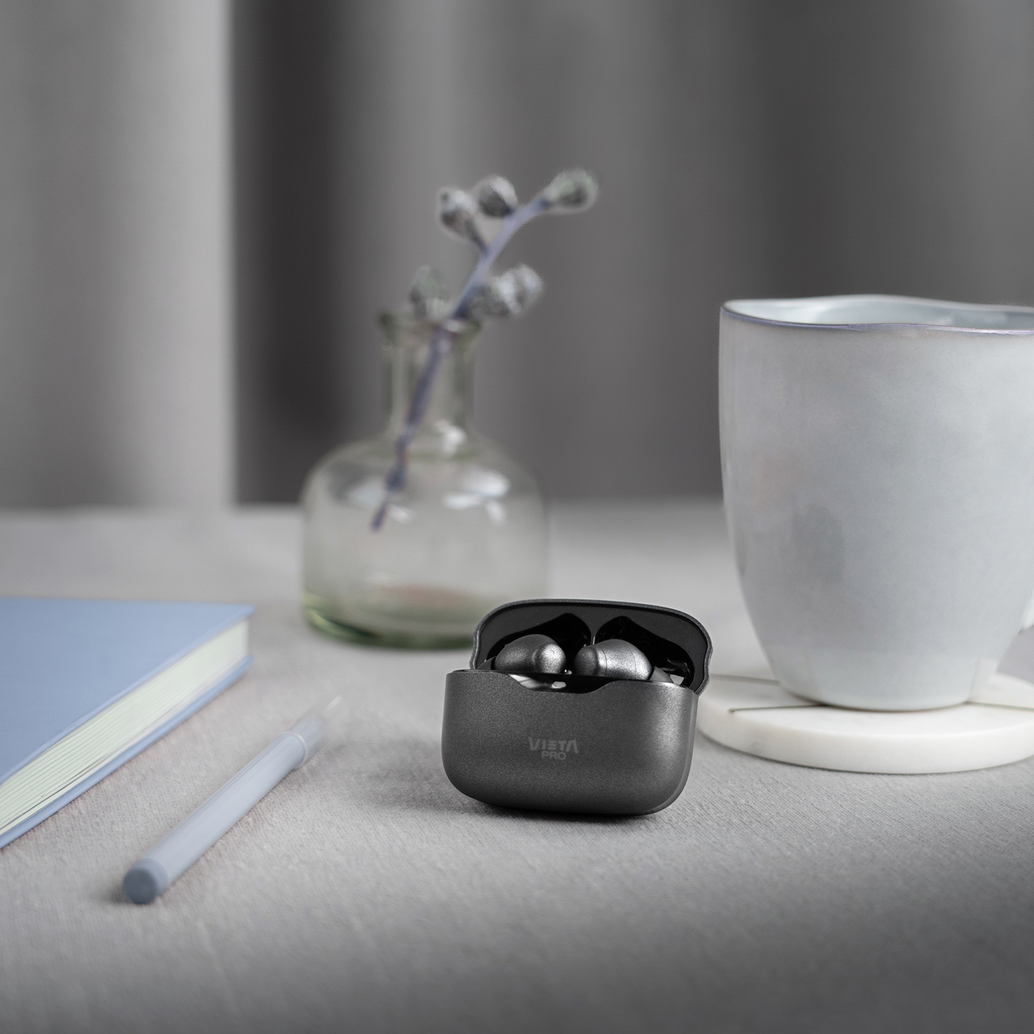 Auriculares en un estuche de carga sobre una mesa, con una taza, un bolígrafo y un jarrón pequeño al fondo.