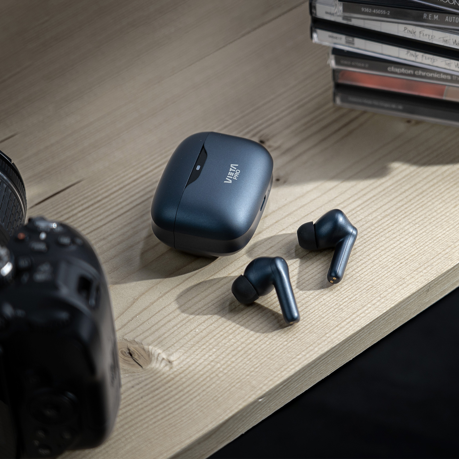 Auriculares inalámbricos azul oscuro y un estuche de carga sobre una mesa de madera, cerca de una cámara.