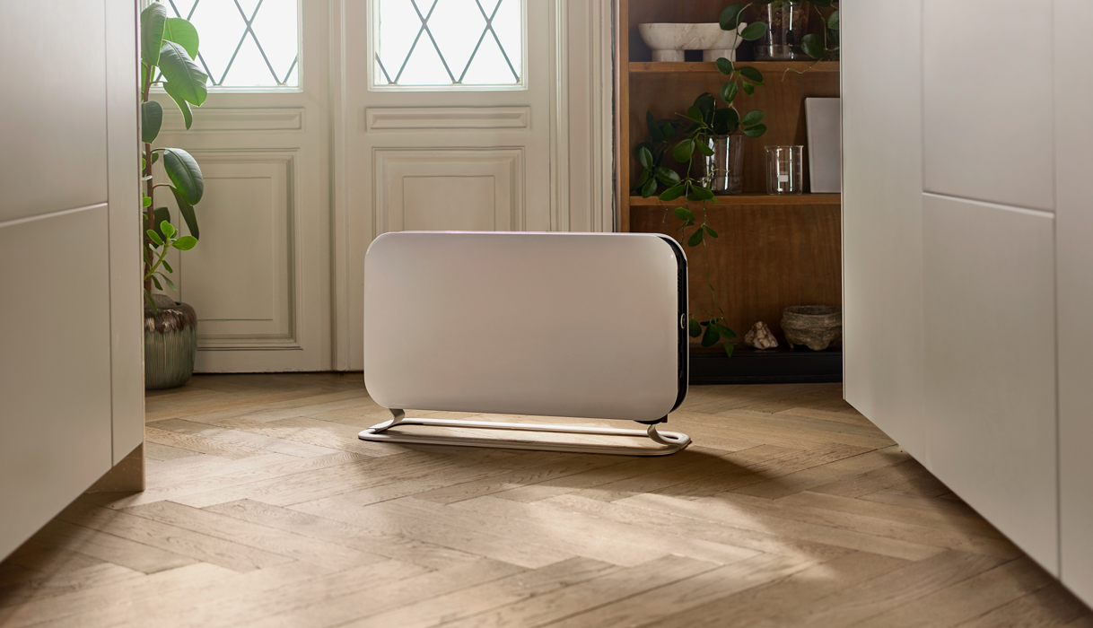 Un termoventilatore elettrico bianco è posizionato su un pavimento in legno vicino a una porta. Lo sfondo include scaffali e porte.
