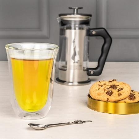 Egy pohár tea, kanál, kávéprés és sütemények az asztalon.