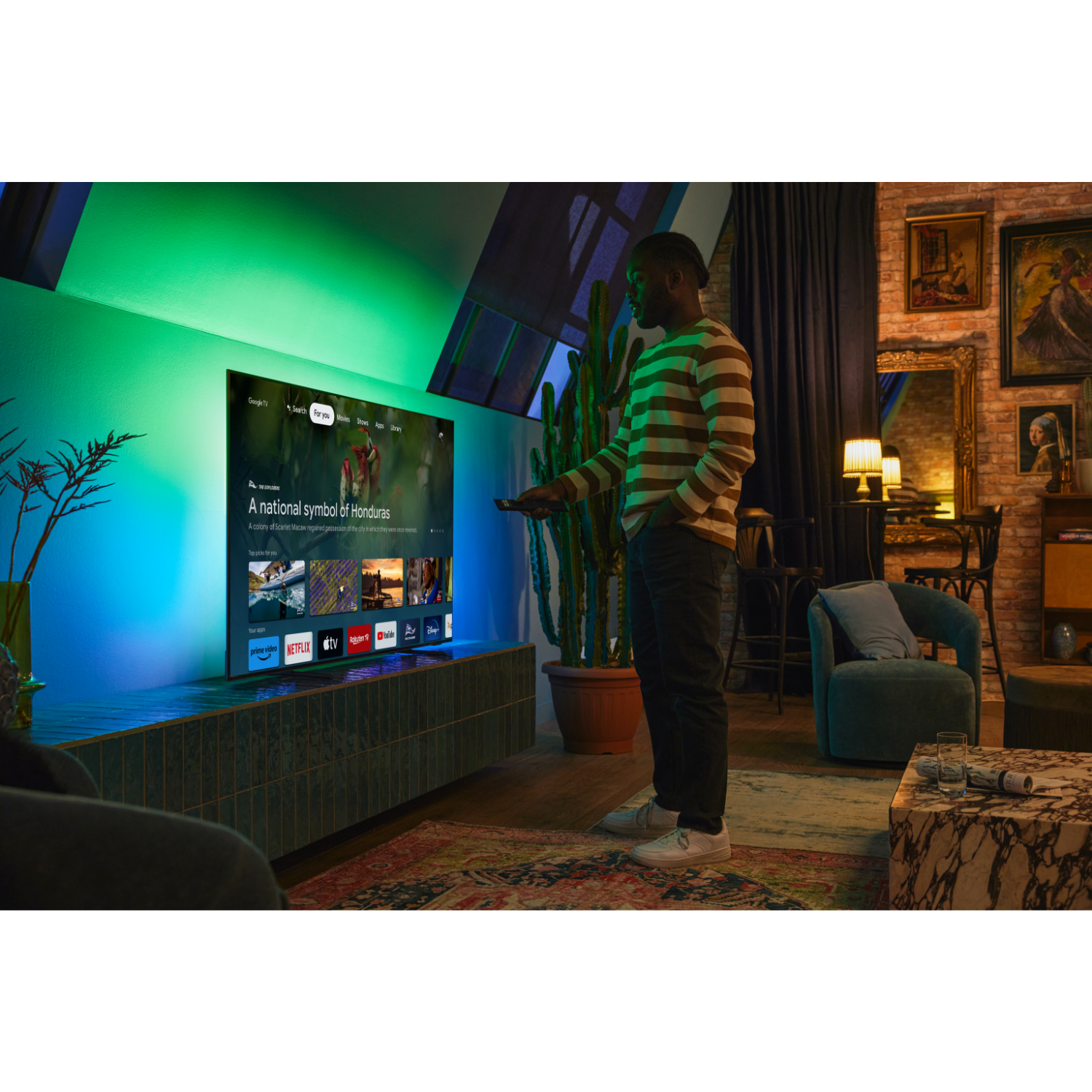 Een man met een gestreept shirt houdt een afstandsbediening vast. Een grote tv staat op de muur. Een cactus staat naast hem.