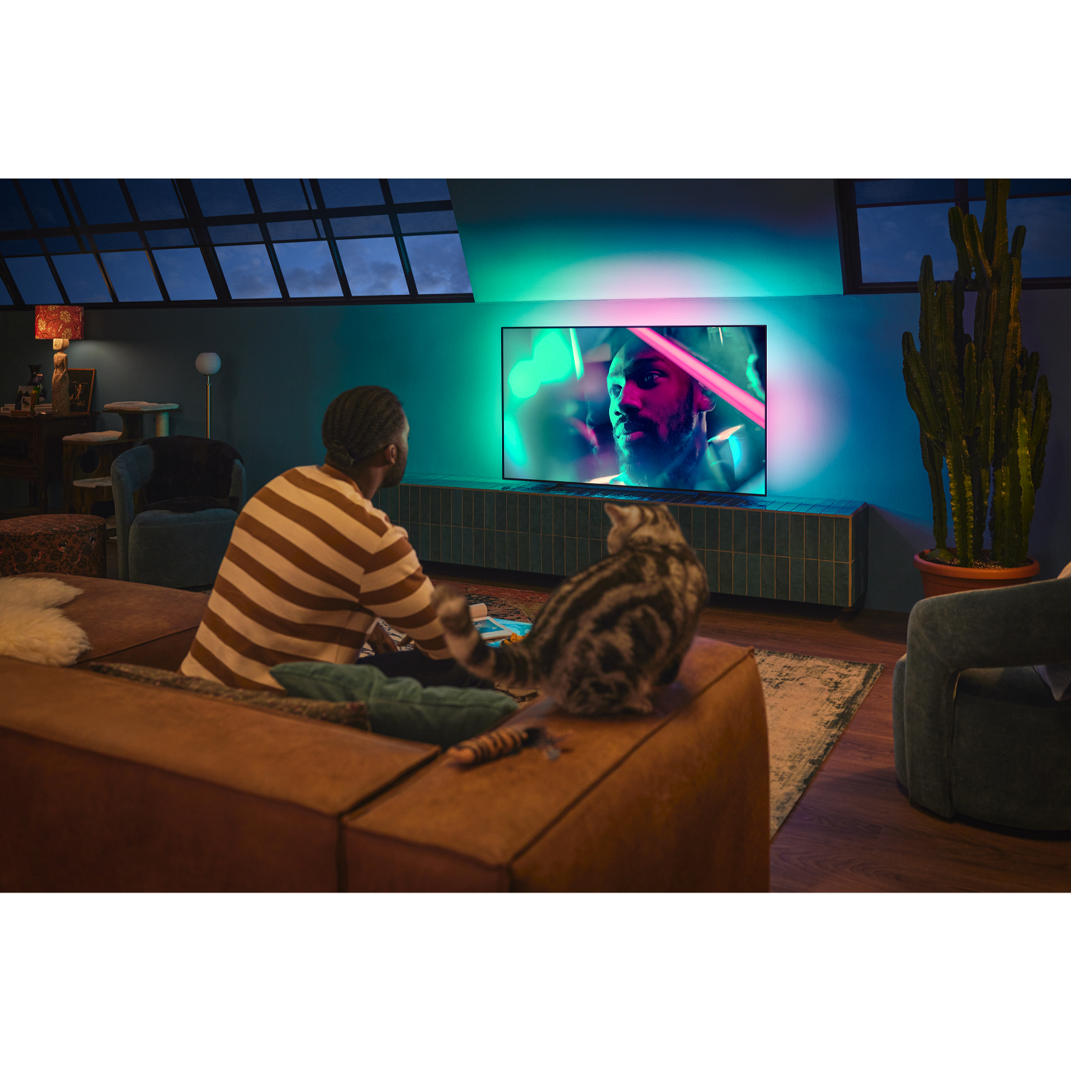 Man kijkt tv met een kat, interieur. De tv staat aan, toont een persoon. Nacht.