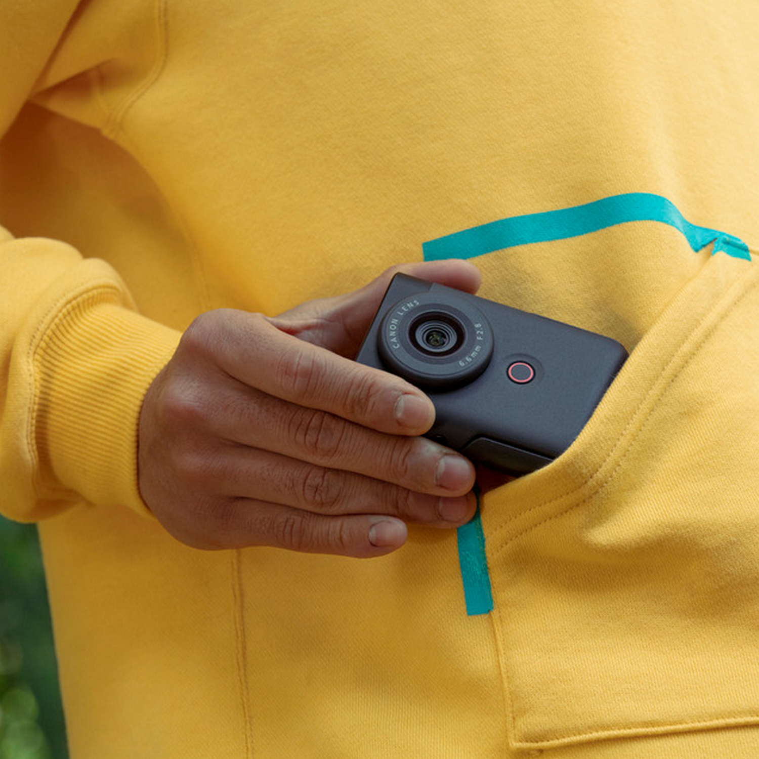 A person's hand holding a small black camera in a yellow pocket. The camera has a lens.