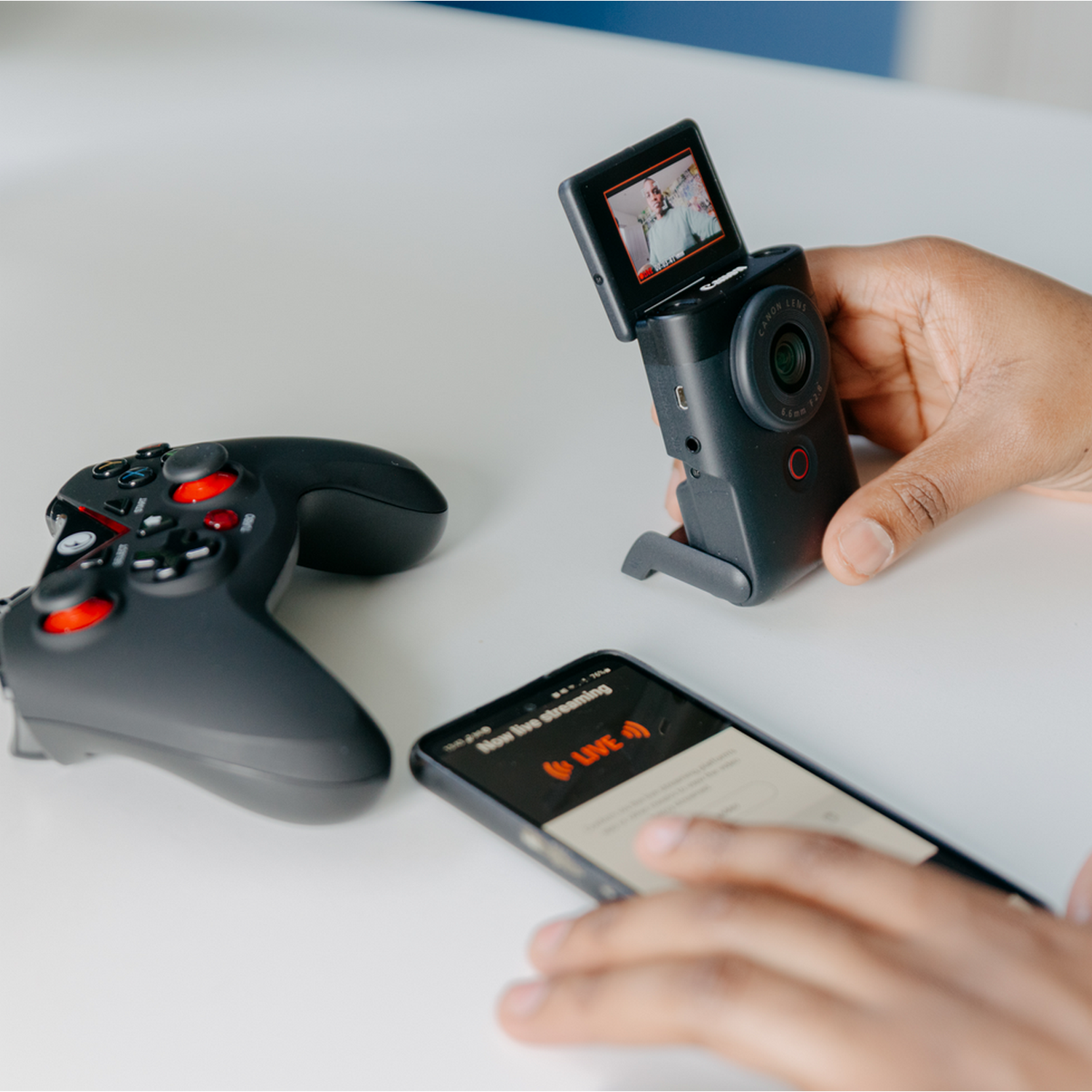 A person's hands with a phone and a camera, with a game controller on the table.