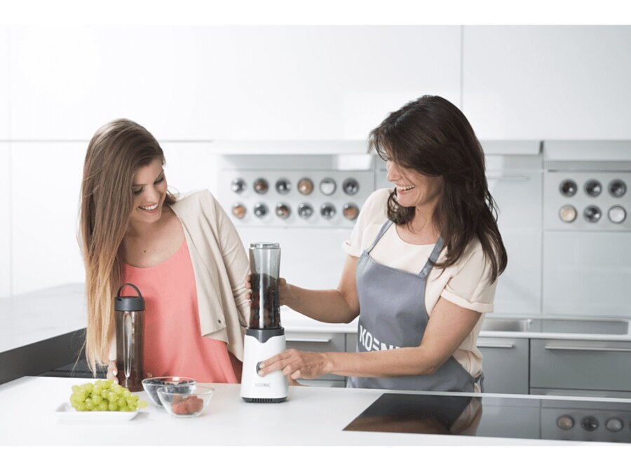 Dwie uśmiechnięte kobiety, jedna używa blendera, otoczenie kuchni.