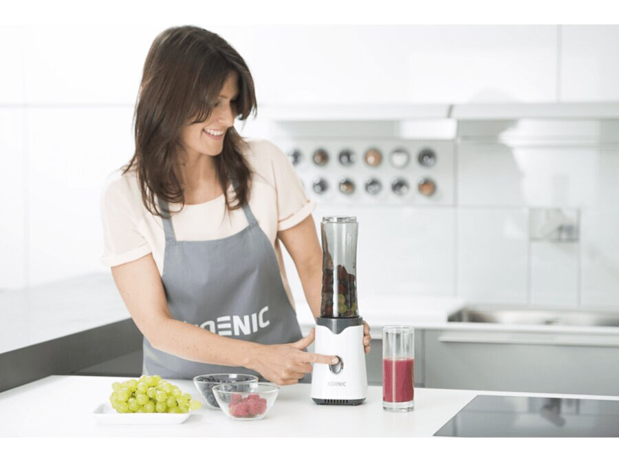 Kobieta uśmiecha się i naciska przycisk na białym i szarym blenderze. Są owoce.