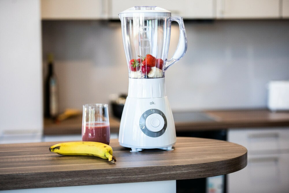 Biały blender z truskawkami i bananem, obok koktajlu i banana na stole.