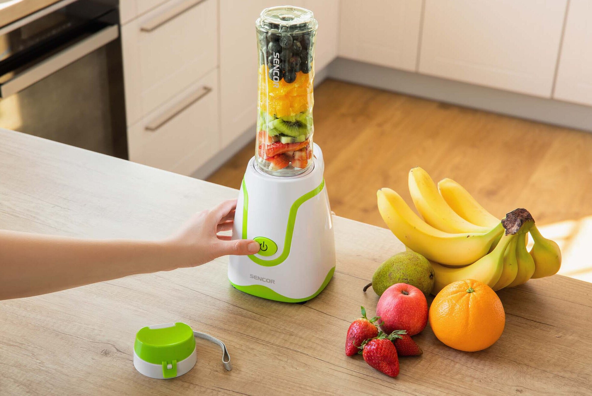 Blender z warstwowymi owocami, ręka naciskająca przycisk, banany i różne owoce na stole.