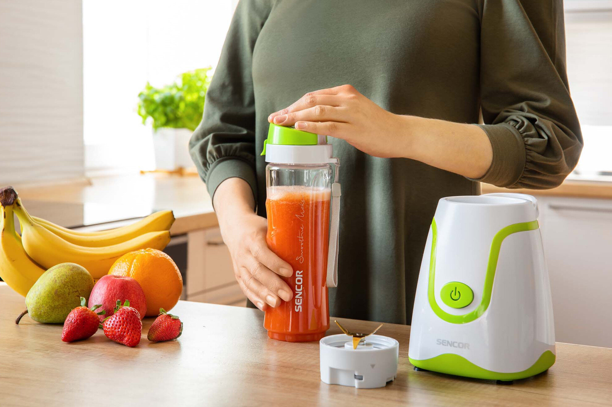 Osoba trzymająca butelkę z sokiem, z owocami i blenderem w tle.
