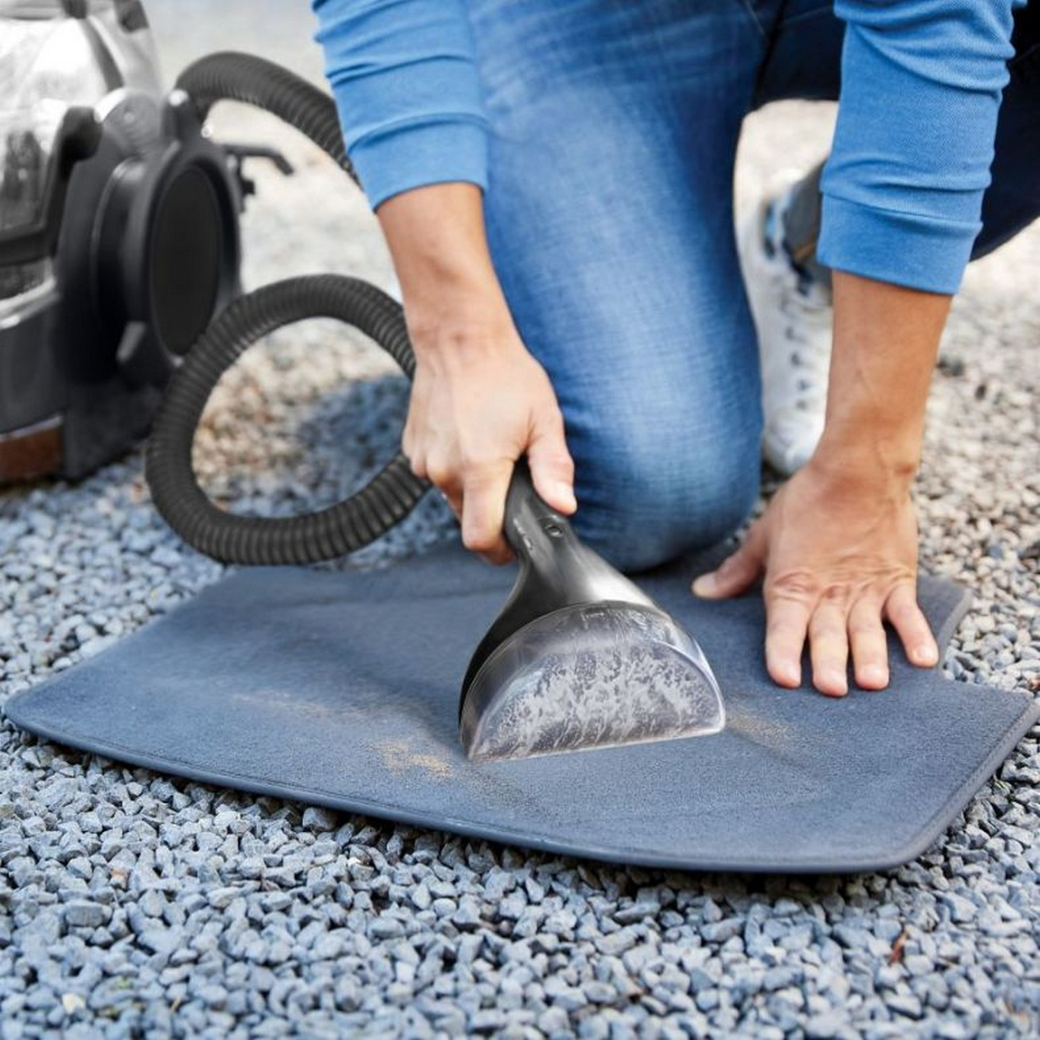 Une personne nettoie un tapis de sol de voiture gris avec un aspirateur sur une surface de gravier.