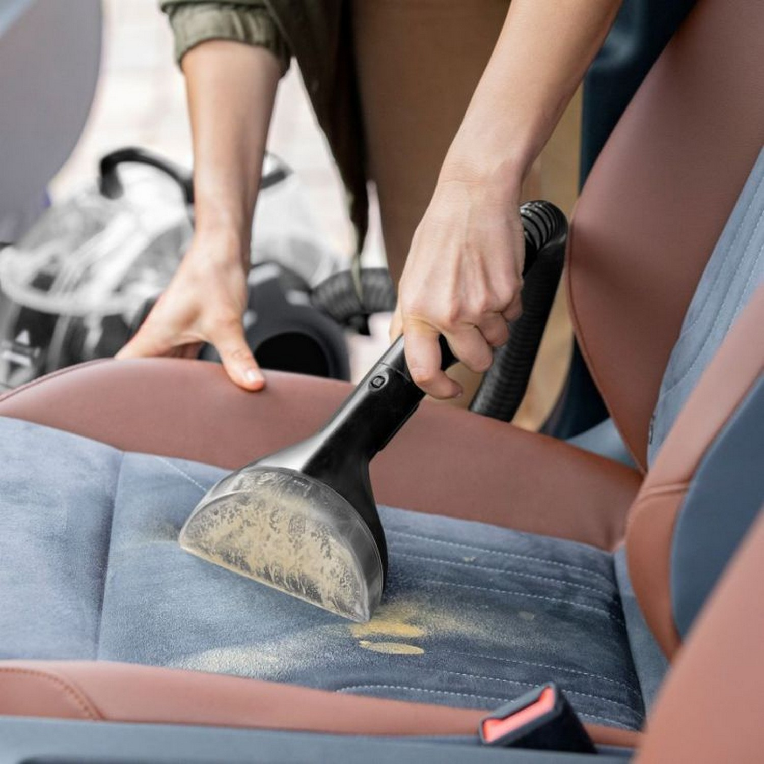 Une main passe l'aspirateur sur un siège de voiture. Le siège est marron et bleu. La tête est noire.