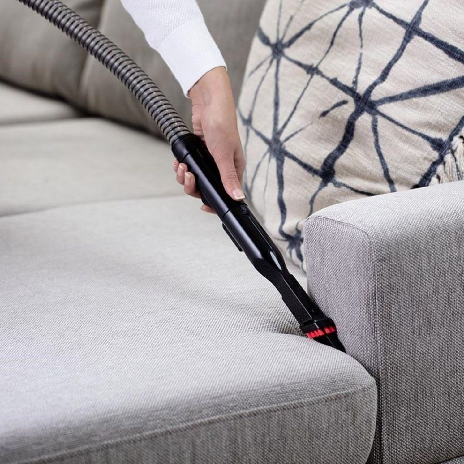 Une main passe l'aspirateur sur un canapé gris. La tête de l'aspirateur nettoie le coin du canapé.