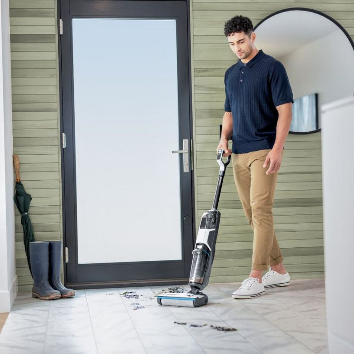 Un homme passe l'aspirateur. Porte, miroir, bottes en arrière-plan. Mur en bois clair.