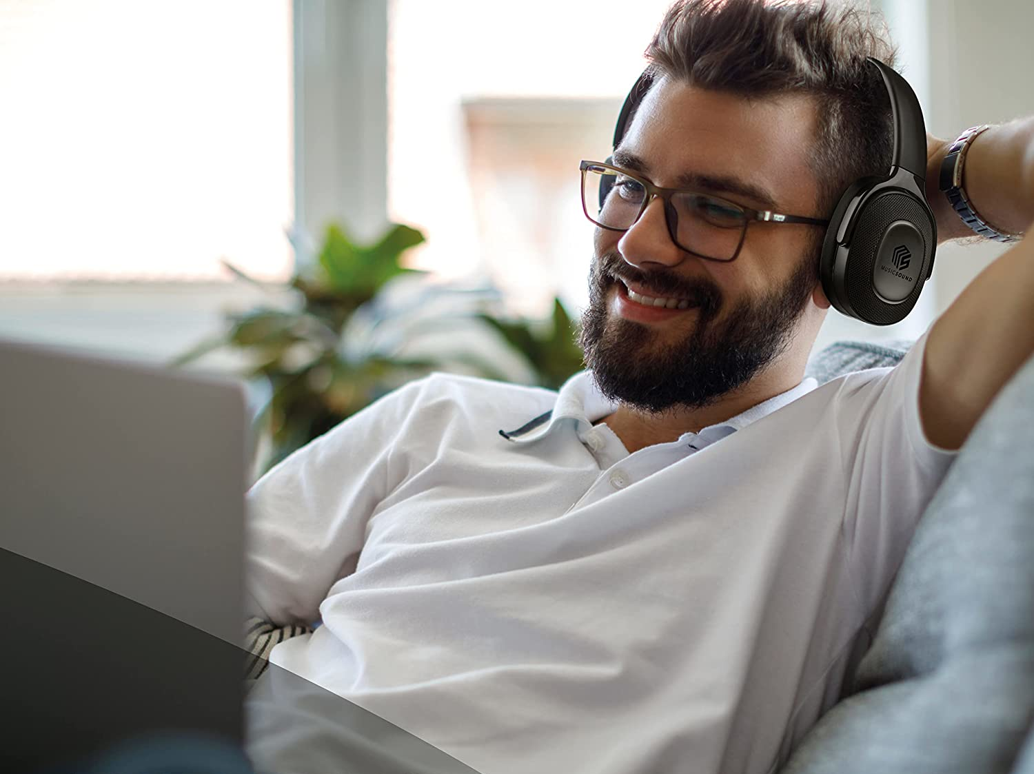 Un homme portant des lunettes et un casque, souriant et regardant un ordinateur portable assis sur un canapé.