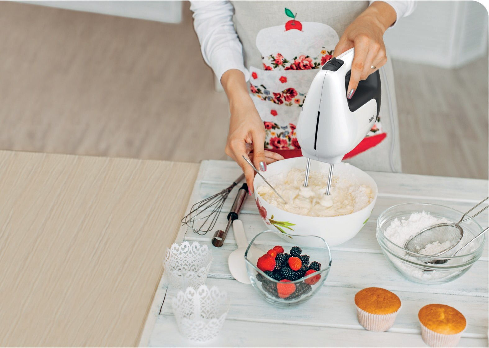 Osoba w fartuchu miesza ciasto w misce mikserem ręcznym, przed nią różne artykuły do pieczenia.