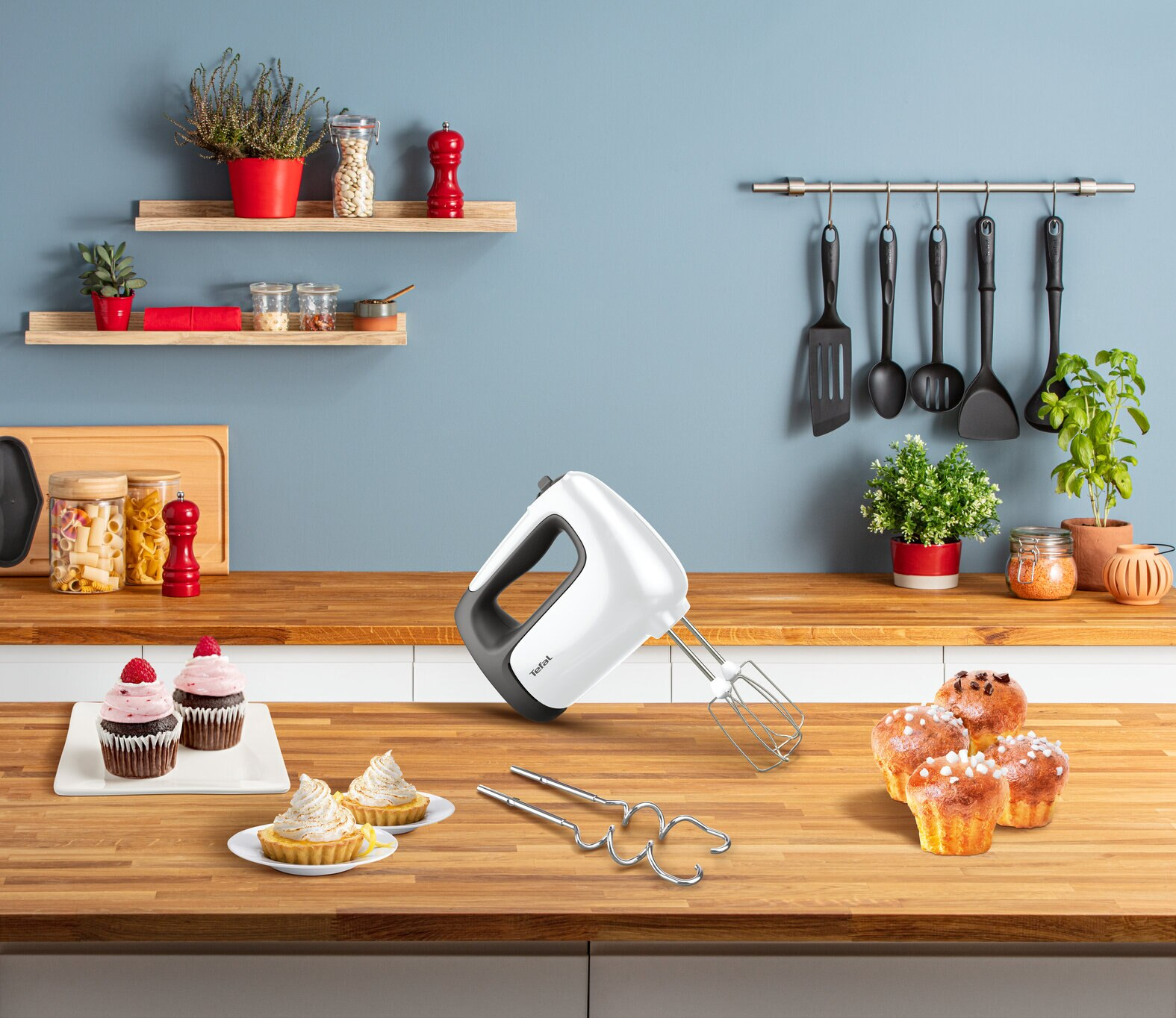 Biały mikser ręczny nad drewnianym blatem z babeczkami, tartami i przyborami kuchennymi.
