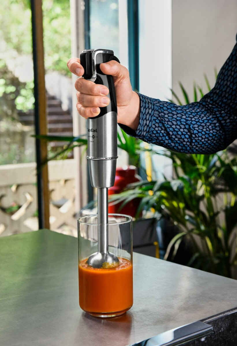 Ręka trzyma srebrny blender zanurzeniowy. Miesza pomarańczowy płyn w szklance.