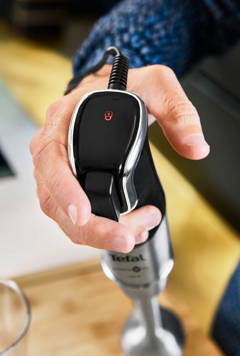 Ręka trzymająca blender ręczny Tefal. Czarny panel sterowania z czerwonym światłem. Rozmyte tło.