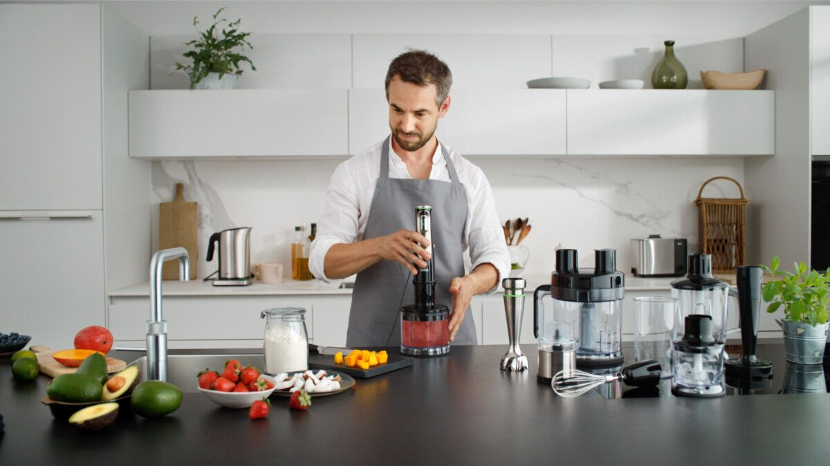 Mężczyzna w fartuchu miksuje czerwoną ciecz blenderem ręcznym. Owoce i akcesoria na blacie.
