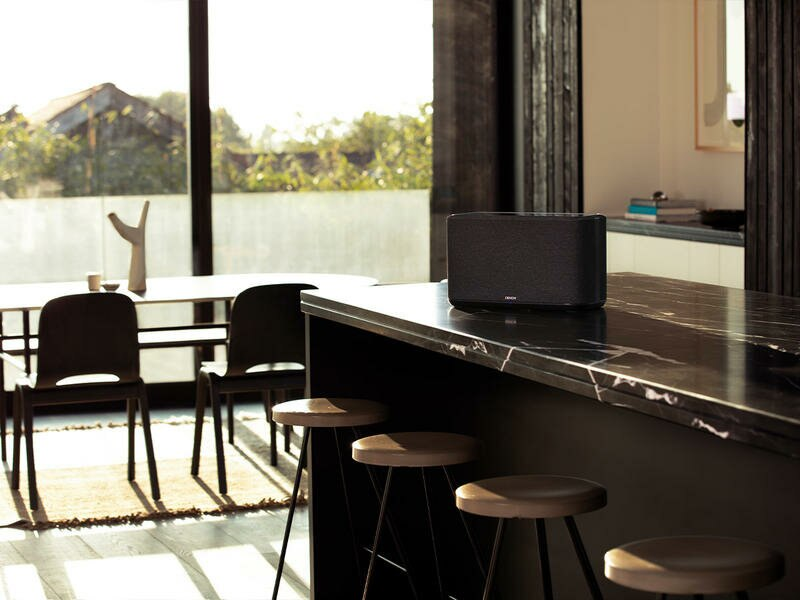 Czarny głośnik na czarnym blacie. Scena we wnętrzu, z krzesłami i oknem w tle.