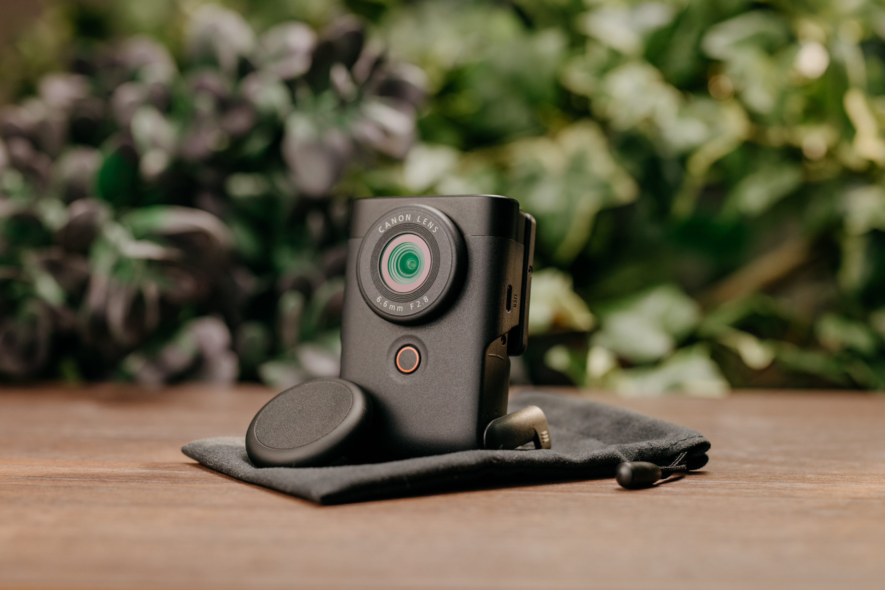 A black Canon camera with lens, mic and accessories on a wooden surface, with green foliage.