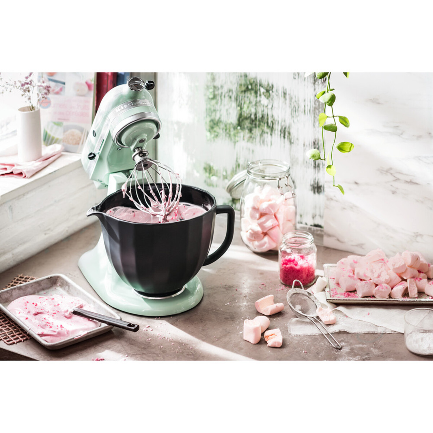 Frullatore KitchenAid con pastella rosa in una ciotola nera. Vasi con marshmallow e vassoio di caramelle rosa.