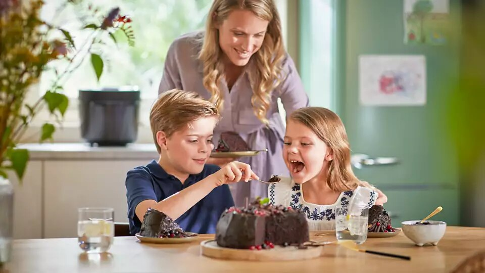 Rodzina, matka i dwoje dzieci, dzielą się ciastem. Dzieci jedzą ciasto, matka patrzy.