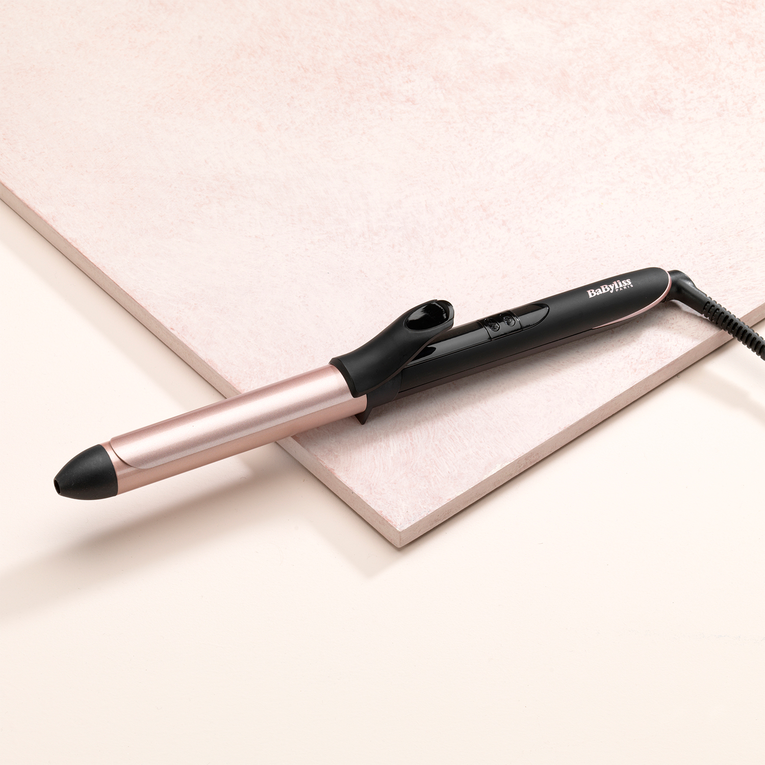 Arricciacapelli, manico nero con fusto oro rosa, posto su due superfici beige rosate e strutturate.
