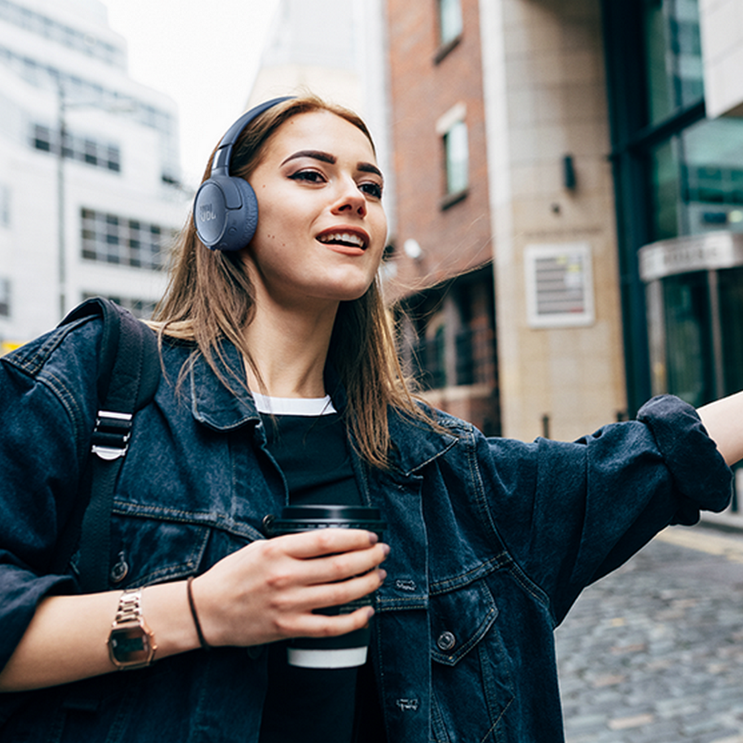 A woman walking on a street with headphones and a coffee cup, looking upwards.
