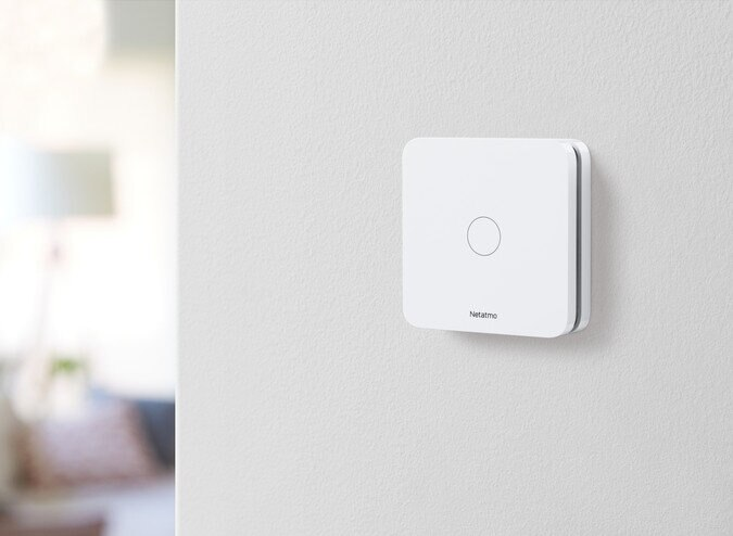 Białe urządzenie smart home Netatmo zamontowane na białej ścianie, z centralnym przyciskiem.