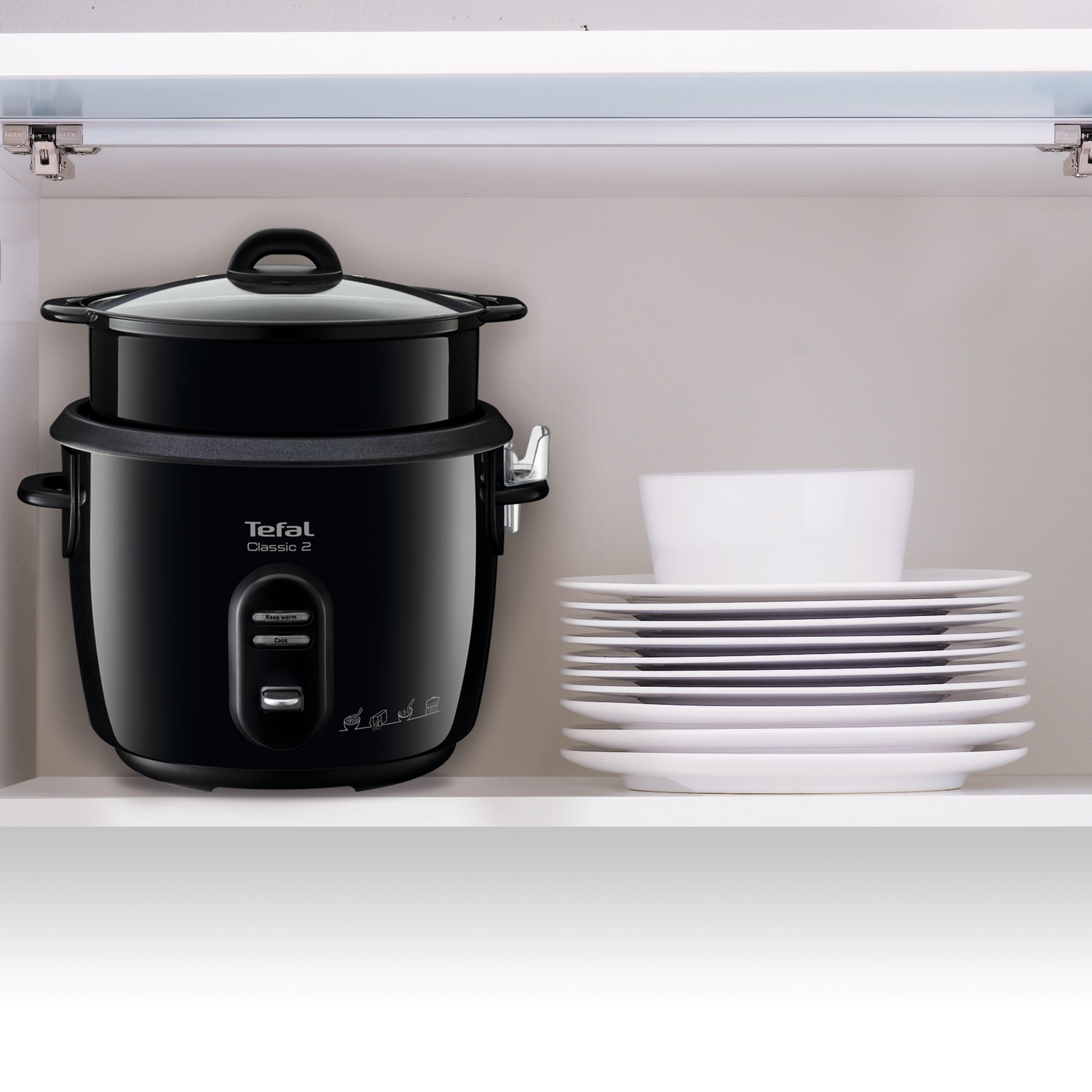 Fekete Tefal rizsfőző egy fehér konyhaszekrényben, fehér tányérokkal a közelben.