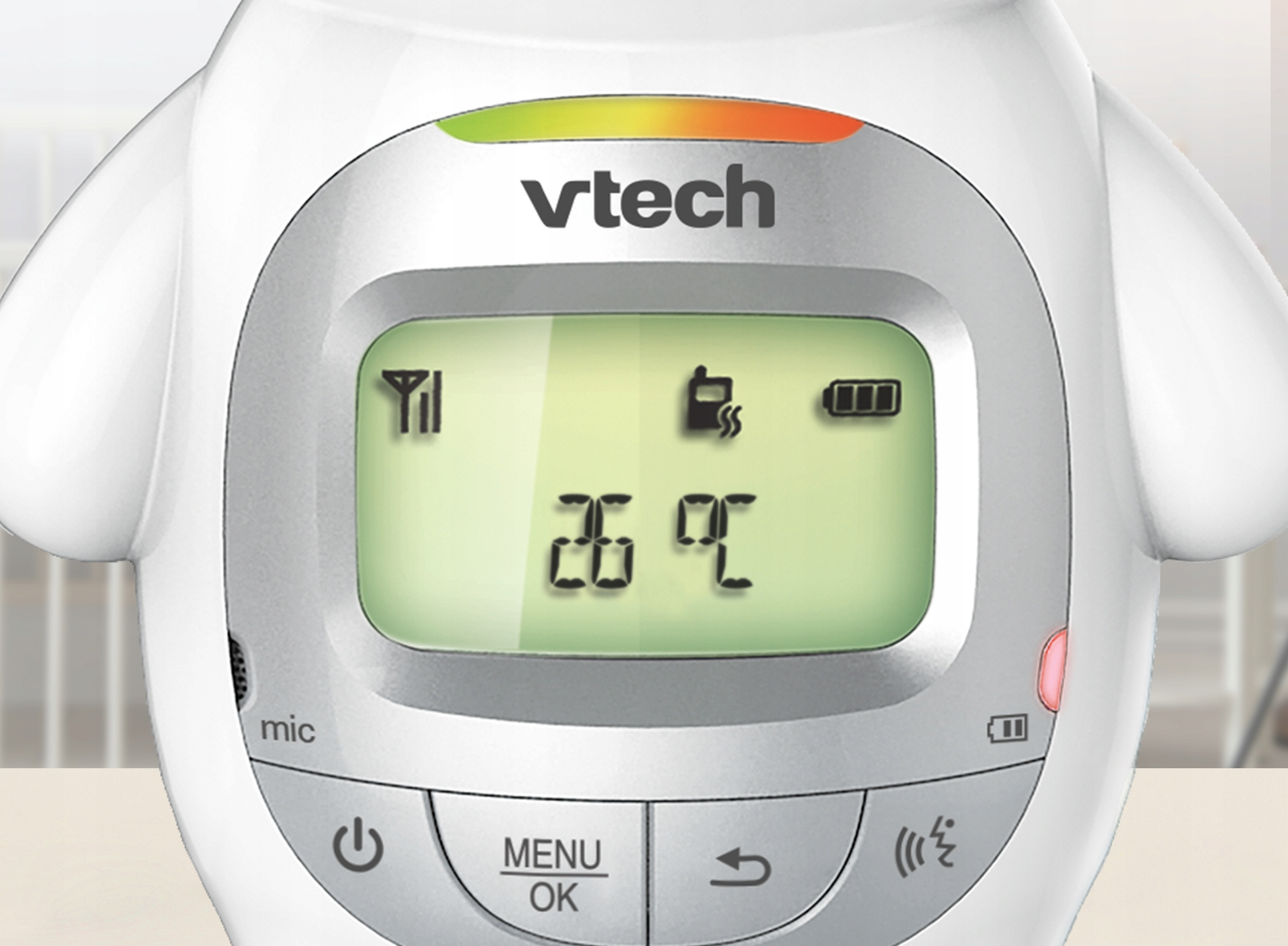 Fehér VTech bébiőrző zöld képernyővel, amely a hőmérsékletet, az akkumulátort és a jelzőket mutatja. Gombok és mikrofon látható.