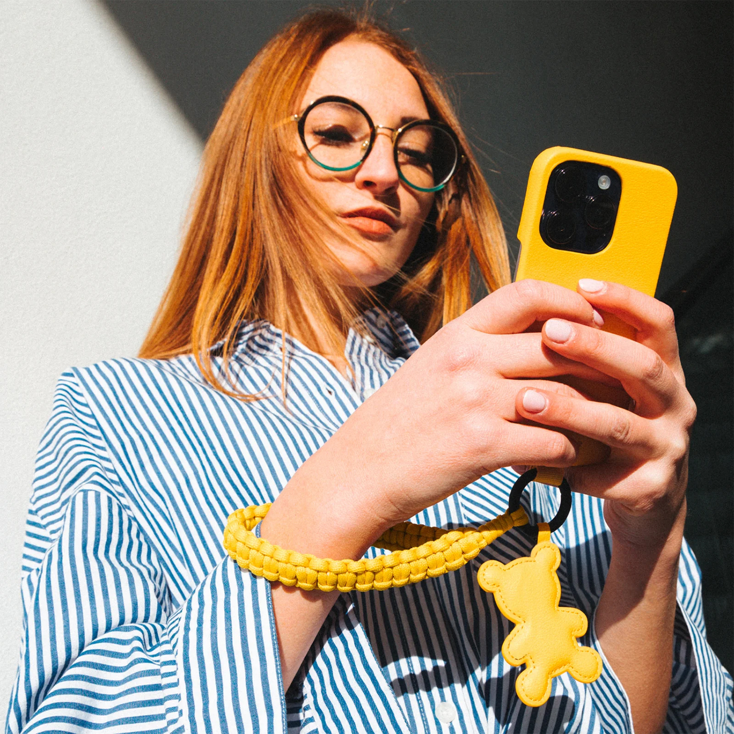 Donna con occhiali che tiene un telefono giallo e un cinturino da polso giallo con un ciondolo a forma di orso.